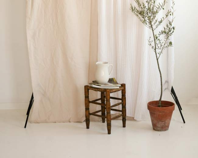 A minimalistic interior scene with a wooden stool, a potted plant, and a pitcher on a small table, separated by two draped fabric panels.