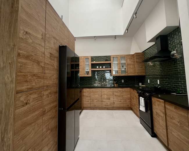 A modern kitchen features wood cabinetry, black appliances, and green tiled backsplash under a high ceiling with a skylight.