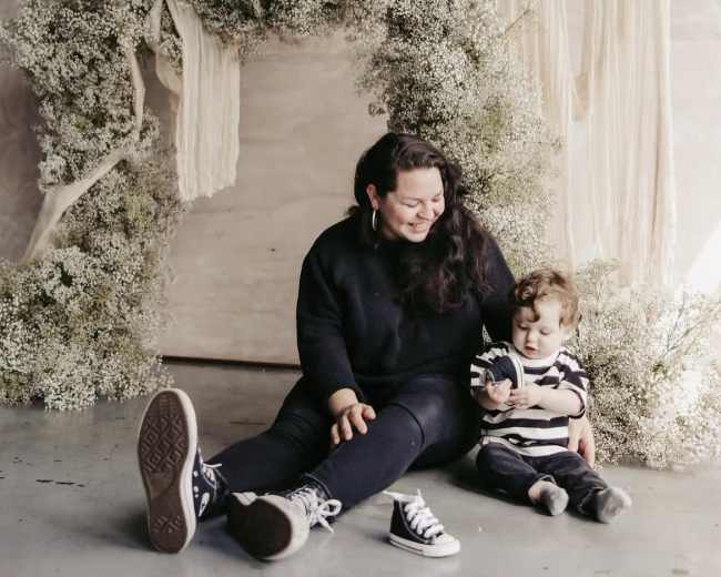 A woman sits on the ground next to a young child in front of a floral backdrop made of greenery and draped fabric.