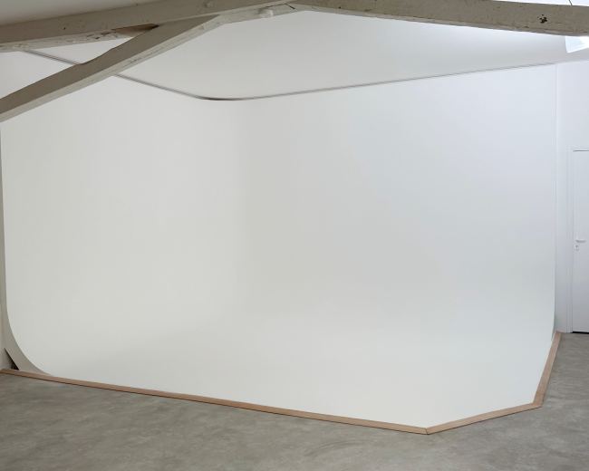 The image shows a minimalistic studio space featuring a seamless white backdrop that curves into the floor, complemented by wooden trim along its edges.