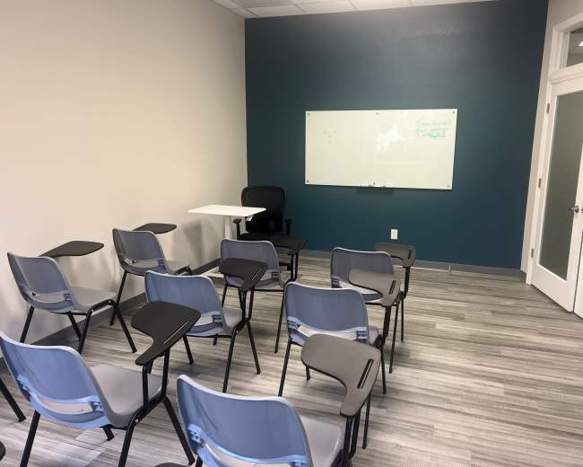 The image shows a small classroom with rows of light blue chairs equipped with writing surfaces, a whiteboard on the wall, and a desk and chair positioned at the front.