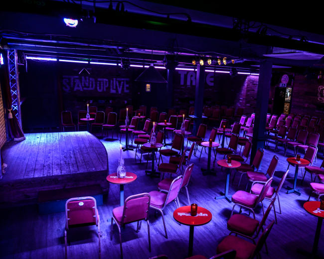 The image shows a dimly lit comedy venue with rows of empty chairs and small round tables facing a small stage.