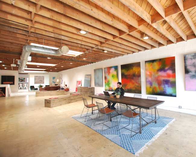 A person works at a table in a spacious room with high wooden ceilings and colorful abstract paintings on the walls.