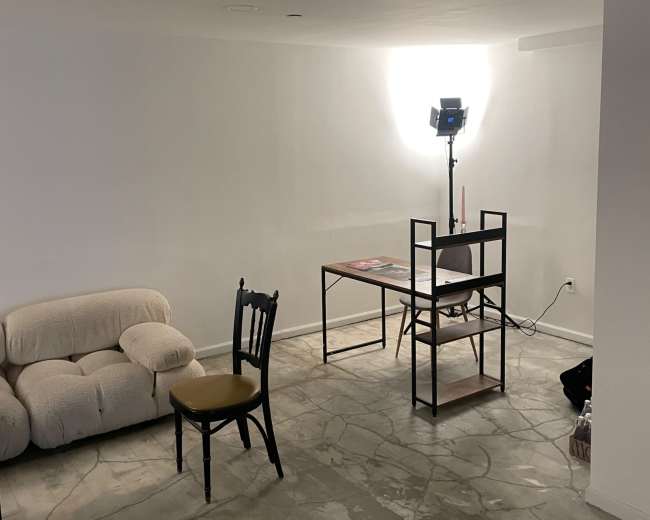 A minimalistic room features a beige couch, a black chair with a yellow cushion, a table with magazines, and a lighting setup in the background.