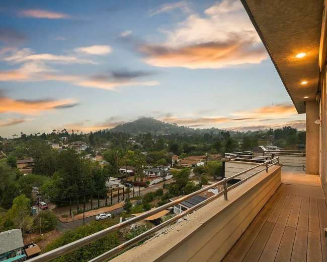 The image shows a balcony view overlooking a residential area with houses and trees, set against a backdrop of hills and a colorful sky at sunset.