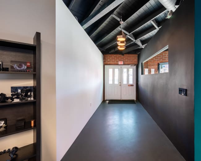 The image shows a narrow hallway with a white wall on one side and a black wall on the other, featuring double doors at the end and exposed brick.