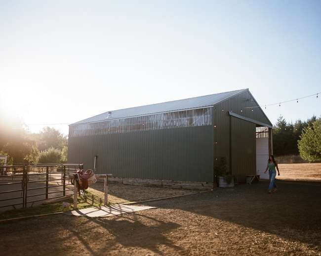 A metal barn stands in a rural landscape under a clear blue sky, with a person walking toward it.