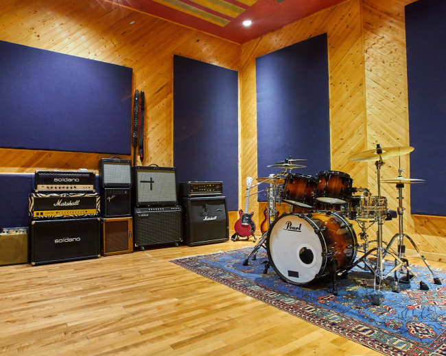 The image shows a music studio featuring a drum set, various amplifiers, and a guitar resting against the wall, all set on a wooden floor.
