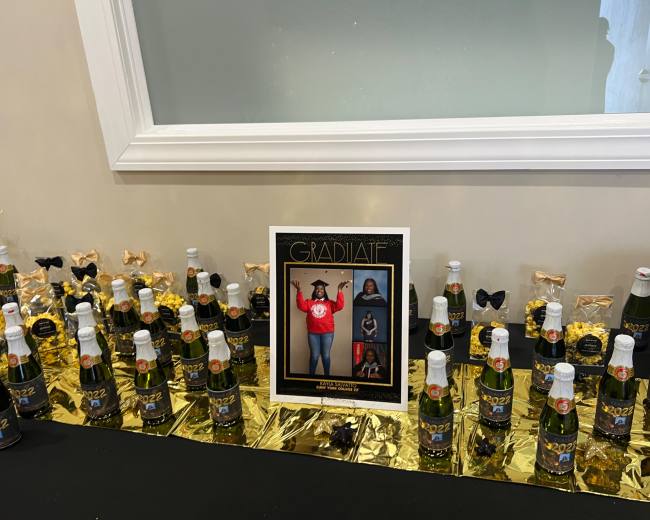 A table is adorned with decorated bottles of sparkling beverage, gold foil, and a framed graduation photo display.