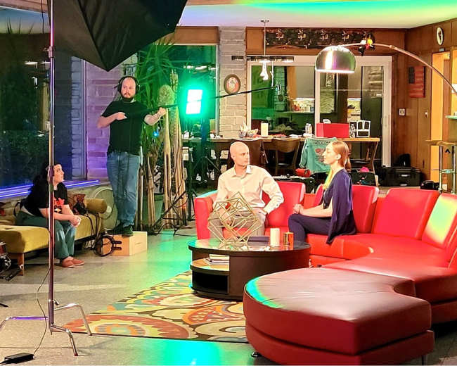A film crew is setting up equipment for an interview scene in a modern indoor space featuring colorful furniture and decorative lighting.
