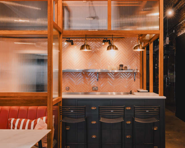 The image shows a modern kitchen area with wood paneling, retro-style cabinets, and a herringbone-patterned backsplash illuminated by pendant lights.