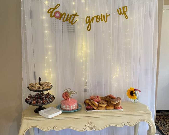 A table displays a variety of donuts and a cake under a decorative banner that reads "donut grow up," framed by a backdrop of white fabric and string lights.