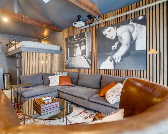 The image shows a modern living area featuring a gray sectional sofa, a glass coffee table, and framed black-and-white photographs on a wood-paneled wall.
