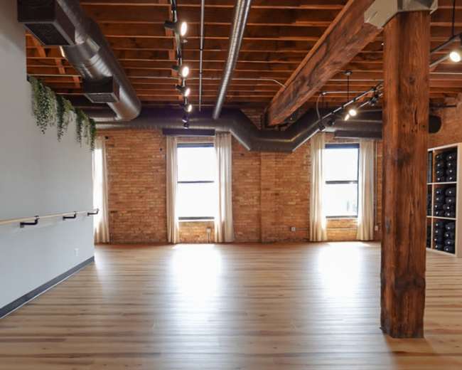 The image shows a spacious interior with hardwood floors, exposed wooden beams, and large windows, creating an open and airy atmosphere.