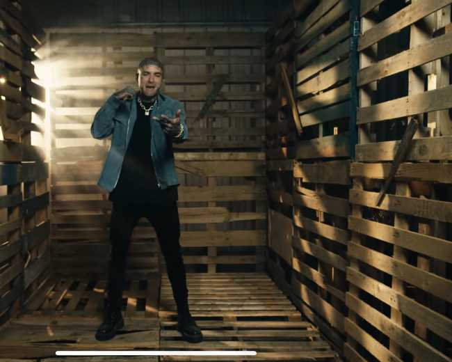 A person stands in a dimly lit wooden structure made of pallets, holding a microphone in a performance stance.