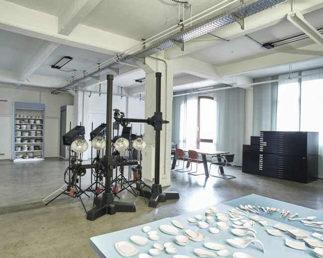 The image shows a spacious studio with equipment for photography, featuring bright lighting setups and neatly arranged tableware displayed on a surface.
