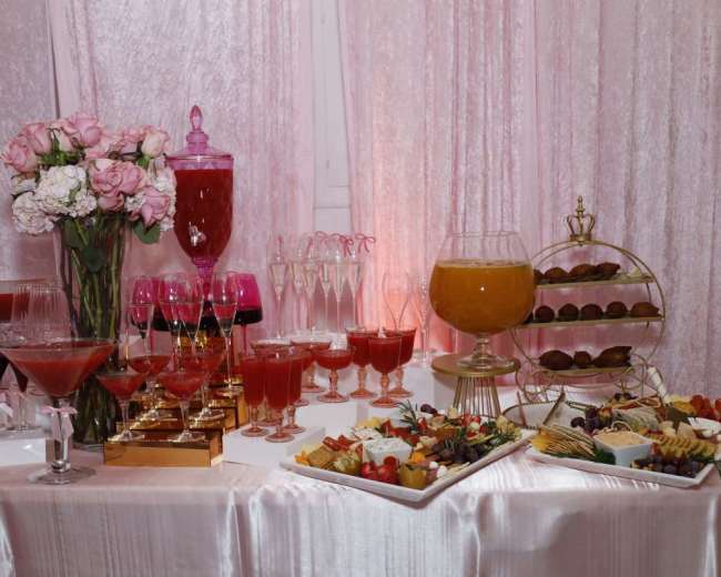 The image depicts a beautifully arranged display of colorful cocktails, floral arrangements, and a variety of appetizers set against a soft pink backdrop.