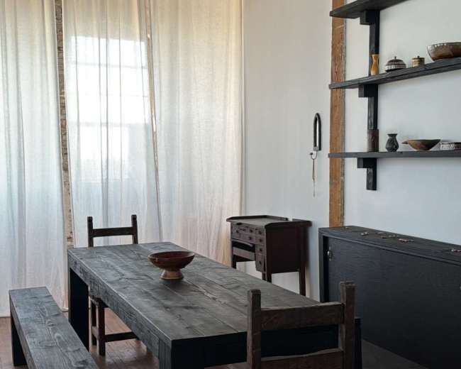 The image shows a minimalist dining area featuring a large black wooden table, a couple of benches, and a small chest of drawers, all set against a backdrop of white curtains and exposed wooden beams.