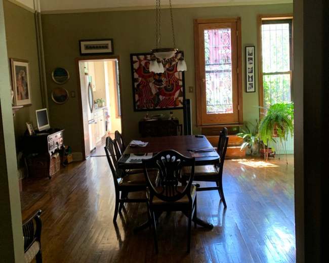 A dining area features a wooden table with chairs, illuminated by a central chandelier, and is bordered by windows and a leafy plant.