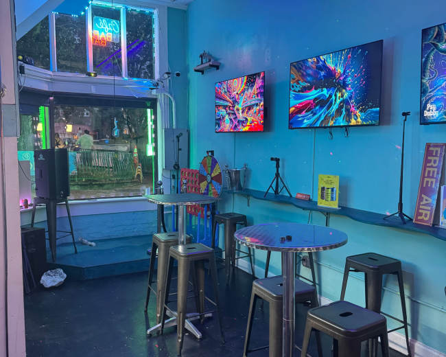 The image shows a brightly lit room with turquoise walls, featuring two large screens displaying colorful visuals, metal bar stools, and tables, along with neon lighting.