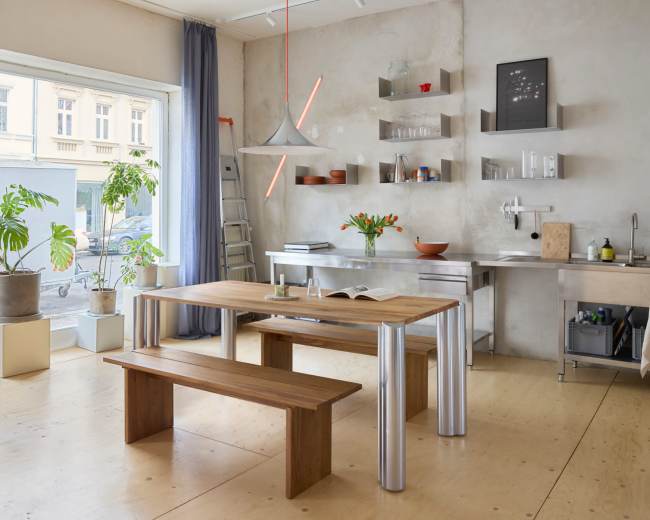 A spacious room features a wooden table with a bench, large window, and a minimalist kitchen area with plants and shelves.