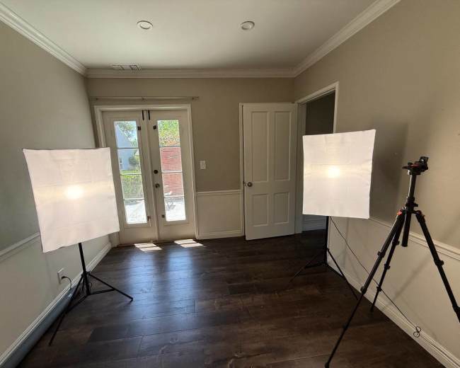 Two softbox lights are positioned on tripods facing a set of double doors in a room with wooden flooring and neutral-colored walls.