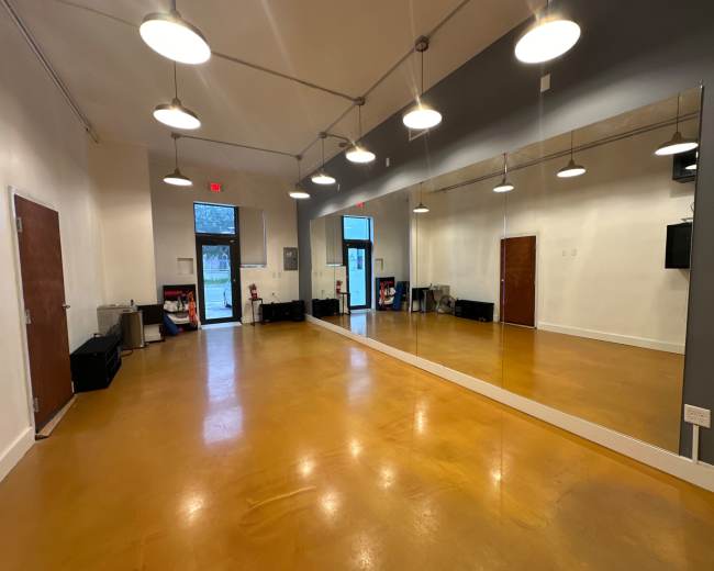 A spacious, well-lit room with a large mirror on one wall, hardwood flooring, and exit doors.