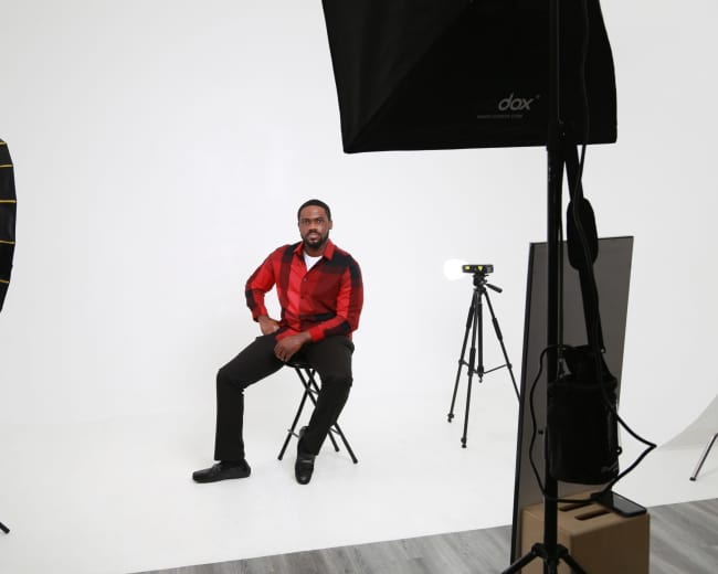 A man sits on a chair in a photography studio, surrounded by studio lighting and a camera on a tripod.