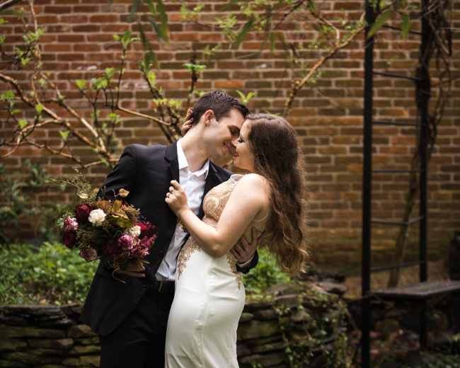 A couple in formal attire embraces in a garden while holding a bouquet, with a brick wall and greenery in the background.