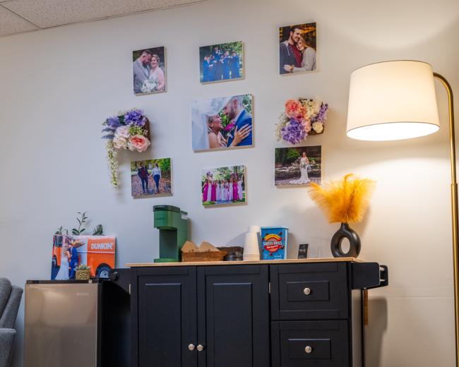 The image shows a black cabinet on wheels with a mini fridge, a lamp, and framed photographs displayed on the wall above.