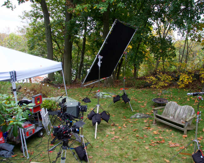 The scene features a film production setup in a wooded backyard, complete with a tent, lighting equipment, and an empty wooden bench.