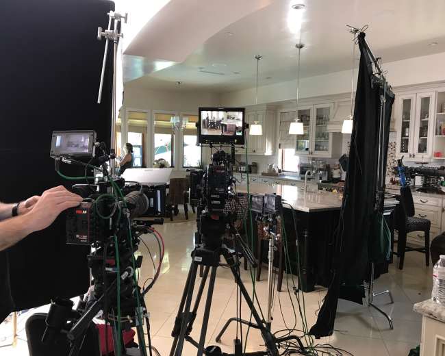 A film crew sets up cameras and equipment in a well-lit kitchen for a shooting scene.