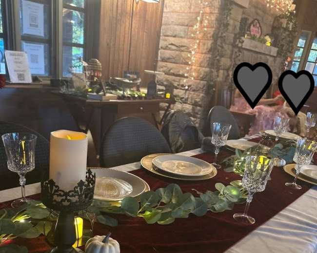 A dining table set with elegant glassware and plates, decorated with green foliage and candles, in a warmly lit room featuring a stone fireplace.