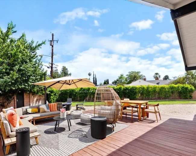The image shows a spacious outdoor patio area with a couch, table, and chairs, surrounded by greenery and under a blue sky with a few clouds.