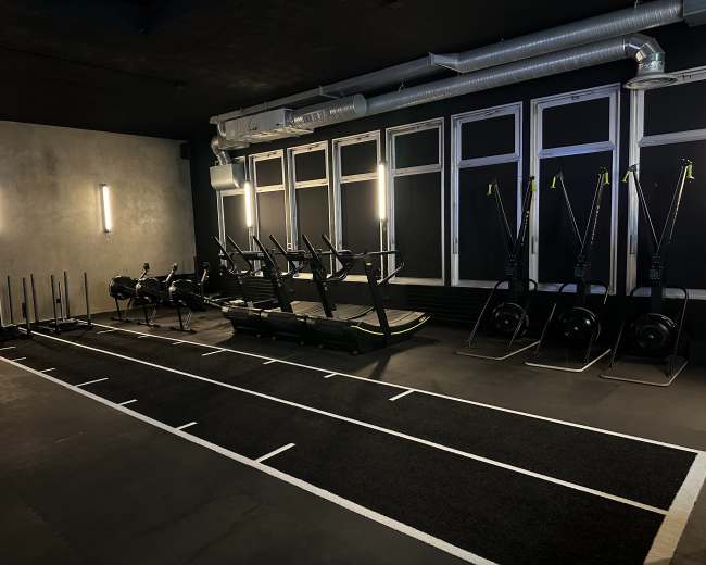 The image shows a modern gym interior featuring exercise equipment such as rowing machines, treadmills, and sleds arranged neatly in a well-lit space.