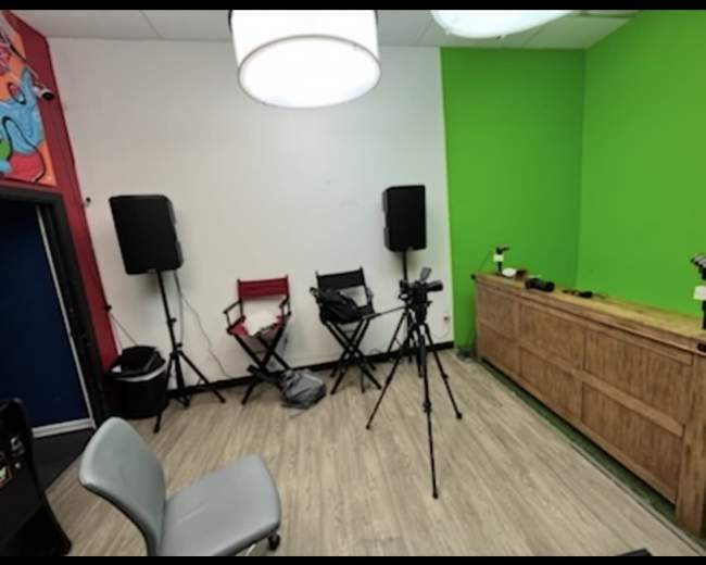 The image depicts a small studio or meeting room featuring a green accent wall, two audio speakers, a wooden storage unit, tripods with cameras, and a couple of folding chairs.