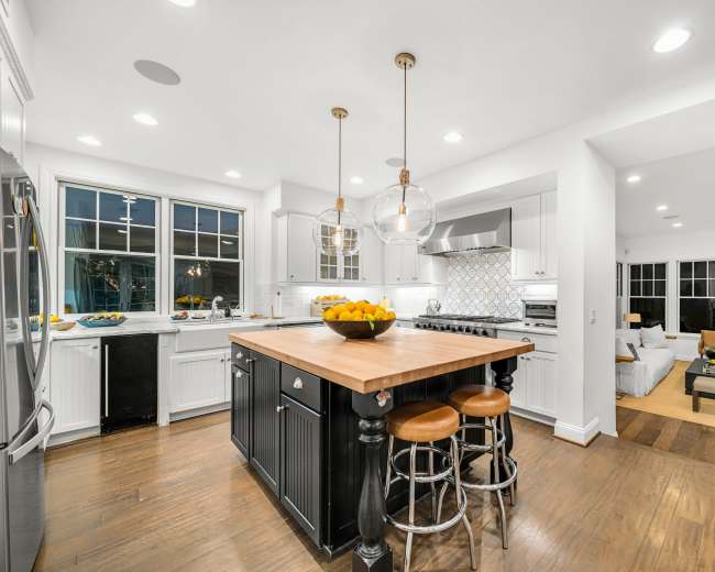 The image shows a modern kitchen featuring a large island with a wooden countertop, three stools, and stainless steel appliances, adjacent to a comfortable living area with a sofa and armchairs.