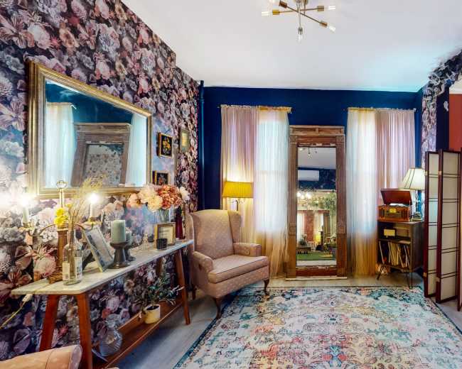A living room decorated with floral wallpaper, featuring a seating area with two chairs and a sofa, a decorative mirror, and a doorway leading to an outdoor space.