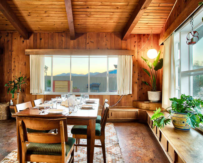 A wooden dining room features a table set for a meal, with large windows displaying mountains in the background.