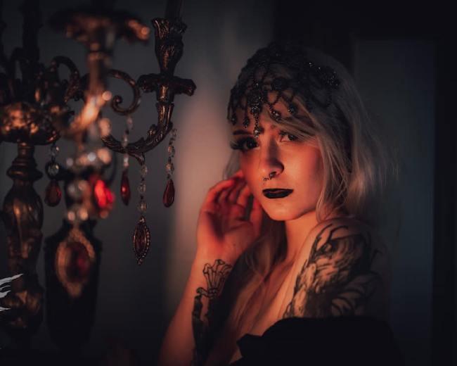 A woman with long hair and tattoos poses beside an ornate chandelier adorned with crystals.