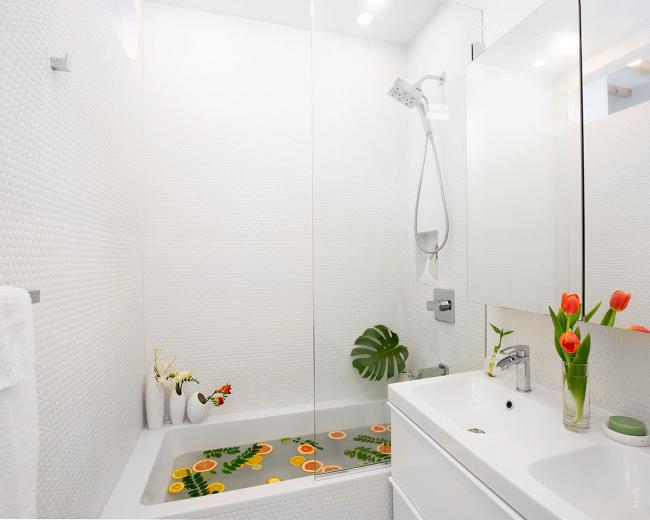 A modern bathroom with a bathtub filled with water and orange slices, surrounded by white tiles and a small flower arrangement.