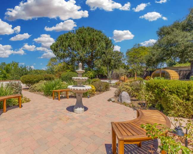 The image shows a landscaped garden area with a central fountain surrounded by benches, shrubs, and trees under a blue sky with scattered clouds.