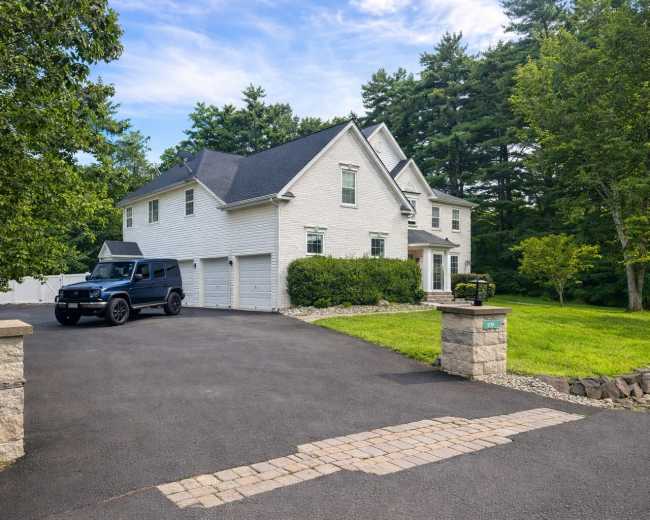 The image shows a large white house with a black SUV parked in the driveway, surrounded by green trees and grass.