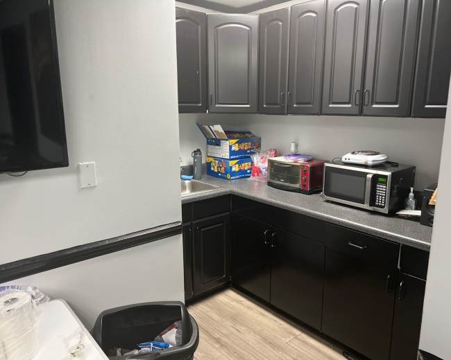The image shows a small kitchen area with black cabinetry, a countertop featuring a microwave, a toaster oven, and various food items, alongside a trash can and a wall-mounted television.