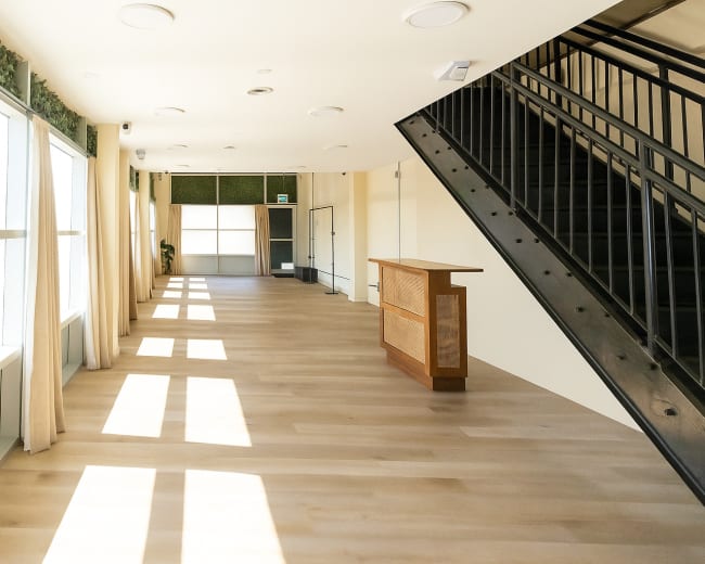 The image shows a bright, spacious interior with large windows, wooden flooring, a reception desk on the right, and a staircase on the left.