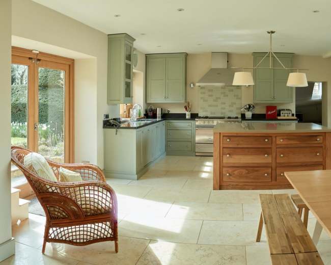 The image depicts a spacious kitchen with modern cabinetry, a large island, a dining table, and glass doors that open to a garden.