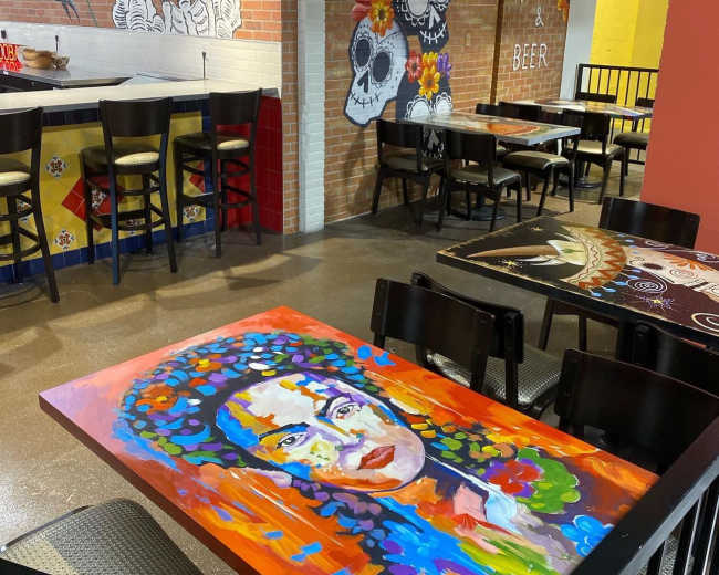 The image shows a vibrant restaurant interior with colorful portraits on the tables and walls, featuring decorative elements inspired by Mexican culture.