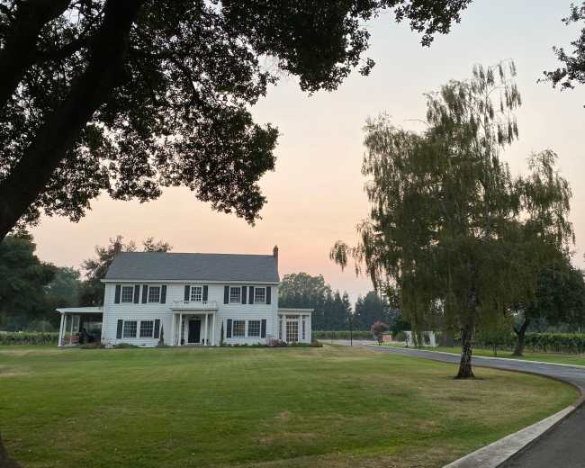 A large white house with a gabled roof stands on a green lawn surrounded by trees, with a winding driveway leading to it.