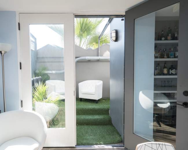 A living space features a pair of glass doors leading to a small outdoor patio with white chairs and artificial grass.