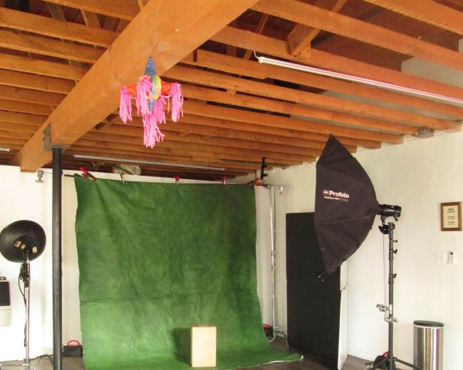 The image shows a photography studio with a green backdrop, a light stand, a photography accessory on the floor, and wooden beams overhead.